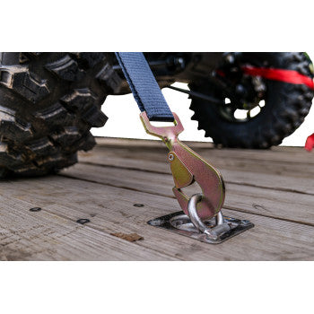 Close-up of TrailBlazer ORV Tie Downs with Carry Bag 4 Pack by Caliber, showing a durable hook fastening a blue polyester strap to a metal anchor on wood, with an offroad vehicle tire in the background—perfect for securing offroad tires.