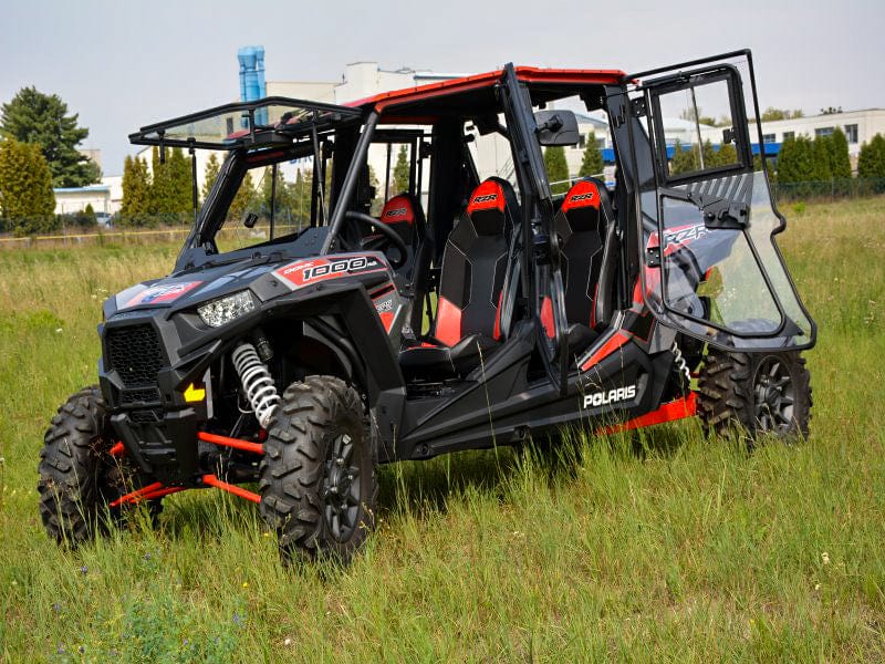 A black and red Polaris UTV with a Full UTV Cab by Open Trail is parked on tall grass. The cab features polycarbonate door panels, rugged tires, visible suspension, four seats, and automotive-quality details. Buildings can be seen in the background.