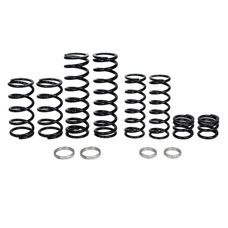 Six black coil springs of different heights from the Polaris Dual Rate Spring Kit by ZBroz are lined up, engineered for improved bottoming resistance. Four silver rings are placed in front on a white background.