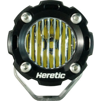 A close-up of the Light Pod Flood by Heretic shows its round, black CNC machined aluminum housing with silver bolts, yellow vertical LED strips, and "Heretic" printed on the front.