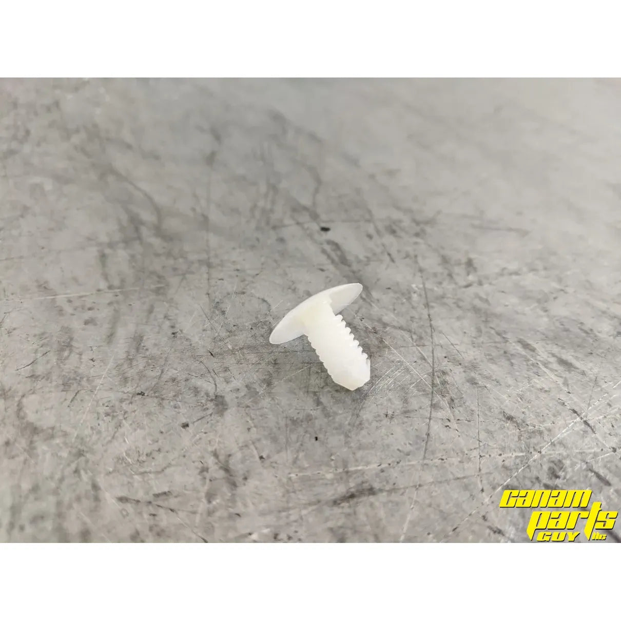 A Trim Button by Can-Am, made of white plastic, rests on a scratched metallic surface. The yellow Can-Am Parts Guy logo in the bottom right corner highlights quality OEM Can-Am parts for your ride.