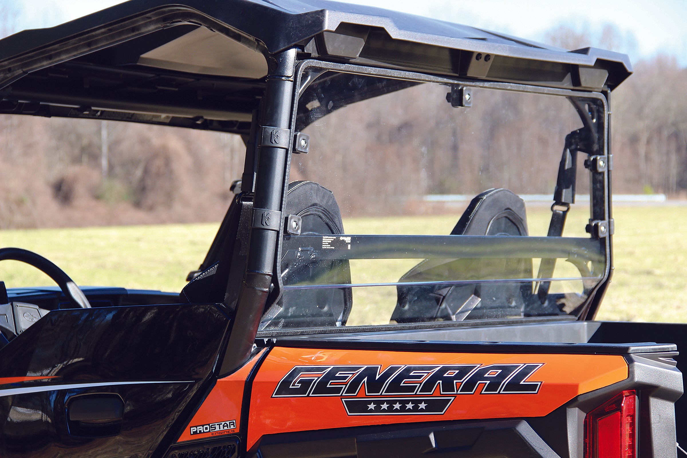 Close-up of a Polaris General 1000, equipped with the Seizmik Rear Hard Dust Panel, showcasing two seats and a clear windshield. The orange off-road utility vehicle is set against a grassy field and trees.