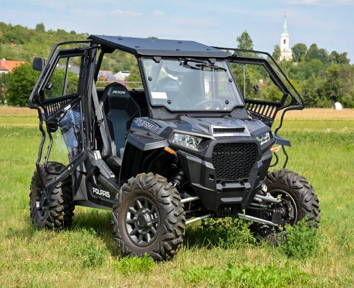 A Full UTV Cab by Open Trail, featuring protective bars and polycarbonate door panels, is parked on grassy land with trees and a church steeple in the background under a blue sky.