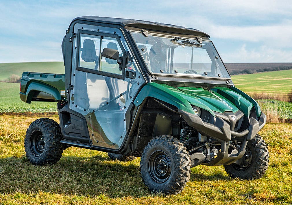 A Full UTV Cab by Open Trail with safety glass and polycarbonate door panels is parked on grass in a rural field, offering great visibility amid rolling hills and a blue sky.