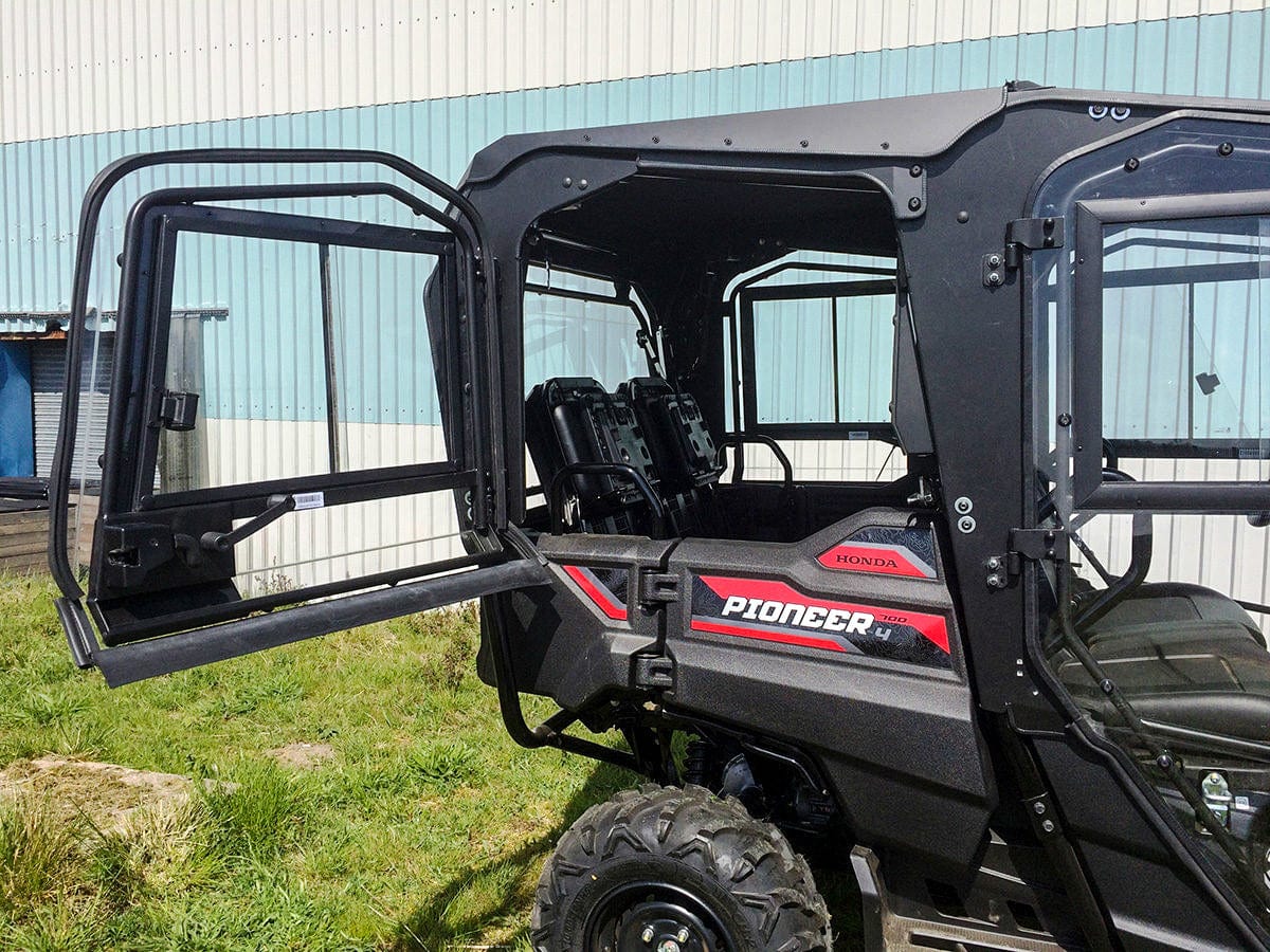 A UTV with the UTV Cab Rear Roof/Back Pane W/Rear Doors by Open Trail is parked on grass near a large industrial building with blue and white metal siding. Its rear doors are open.