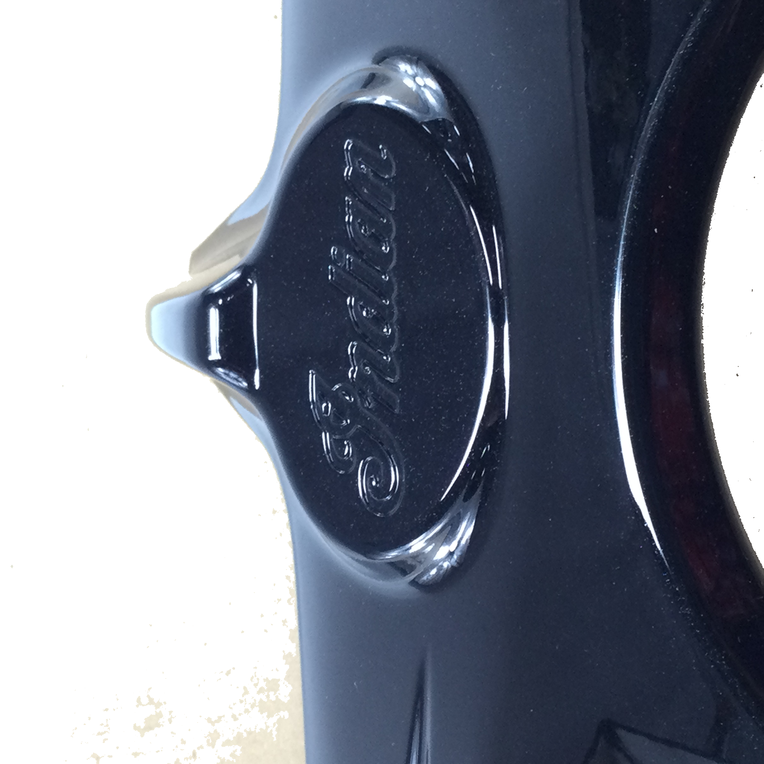 Close-up of a shiny "Headlight Bezel Paint Black Pearl by Polaris" for Indian motorcycles, featuring “Indian” engraved in cursive on a raised oval. Its lustrous surface reflects light, highlighting authentic OEM Indian Polaris parts quality.