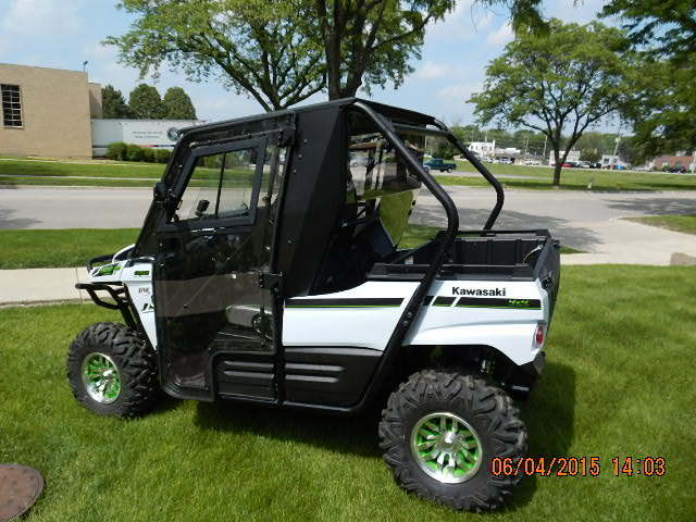 A Full UTV Cab by Open Trail, featuring black and white design with green wheels and durable polycarbonate door panels, is parked on grass near a road, surrounded by trees and buildings, offering enhanced visibility.