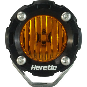 The Light Pod Amber Flood by Heretic features a round LED with an amber lens, black housing, visible corner screws, and “Heretic” in white at the base. Its aircraft-grade aluminum build gives it a rugged, industrial look.