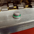 Small metallic hex socket plug with green O-ring gasket on a gray metal surface.
