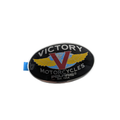 Oval black metal fuel tank badge with silver and gold winged V logo and Victory Motorcycles text.