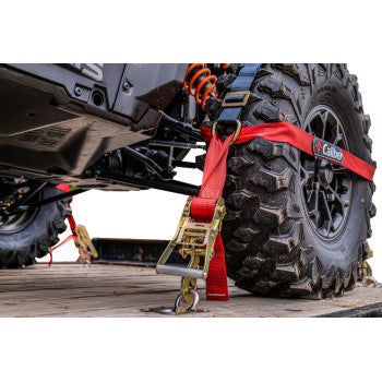 Close-up of a rear tire secured with TrailBlazer ORV Tie Downs with Carry Bag 2 Pack by Caliber—adjustable polyester straps and metal hooks ensure safe offroad vehicle transport. Tire and suspension are visible.