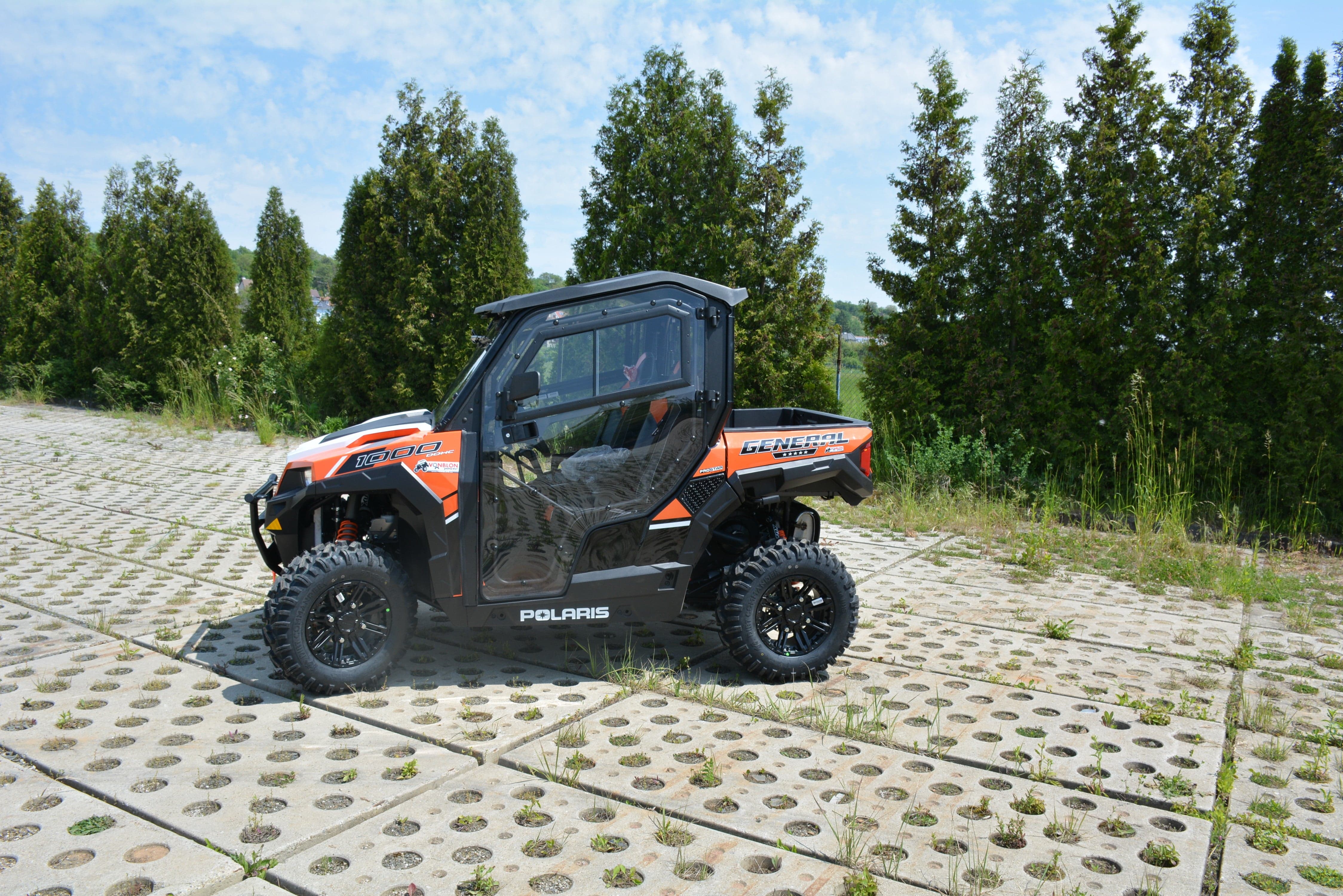 A vibrant orange Polaris equipped with the Full UTV Cab by Open Trail, featuring polycarbonate door panels, is parked on a stone-paved spot among green trees under a partly cloudy sky for optimal visibility on every adventure.