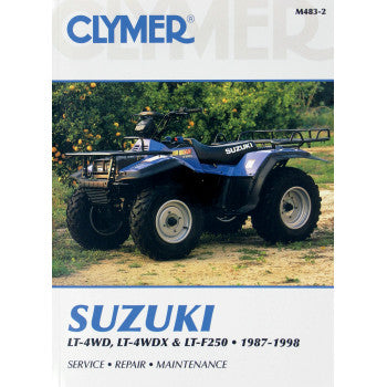 A Suzuki ATV sits on grass by trees. The cover reads: Suzuki King Quad / Quad Runner Manual 250 by Clymer, 1987-1998. Service manuals for maintenance and repair are included.