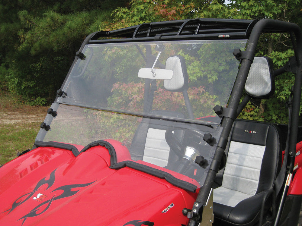 A Polaris Ranger Pro-Fit Full Size Folding Versa Windshield by Seizmik, with custom designed clamps and multiple black fasteners, is securely attached to a red off-road vehicle parked outdoors near green foliage.