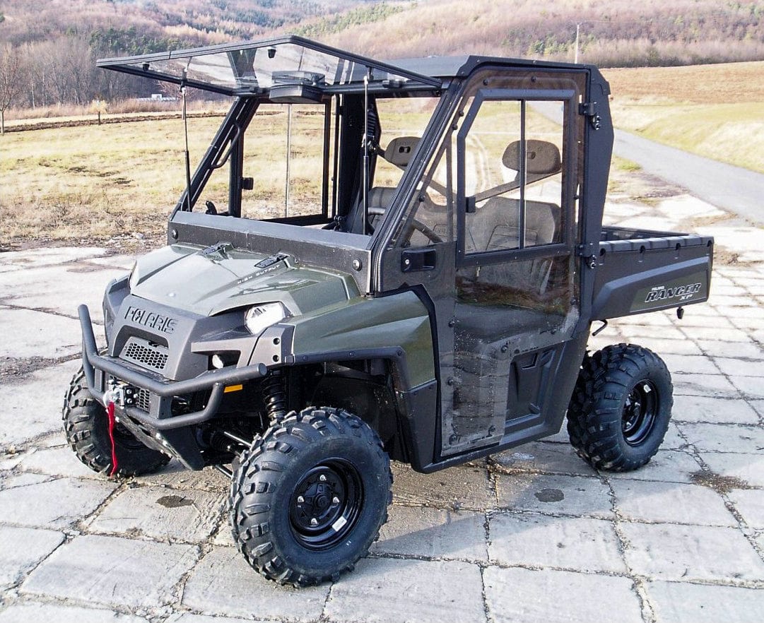 A green Polaris off-road vehicle equipped with the Full UTV Cab by Open Trail, featuring polycarbonate door panels and large tires, is parked on pavement outdoors, with fields and hills in the background.