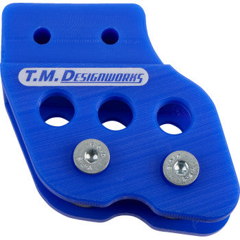 The Honda - Yamaha - Kawasaki - Suzuki Chain Guide Blue by T.M. Designworks features durable blue plastic with three large holes, two metal screws, curved edges, UV protection, and “T.M. DESIGNWORKS” text on its ridged surface.