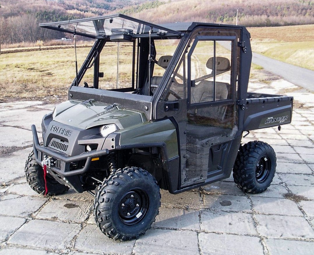 A Full UTV Cab Crew Model by Open Trail with a green exterior and polycarbonate door panels is parked on a stone driveway in a rural landscape with hills and sparse vegetation in the background.