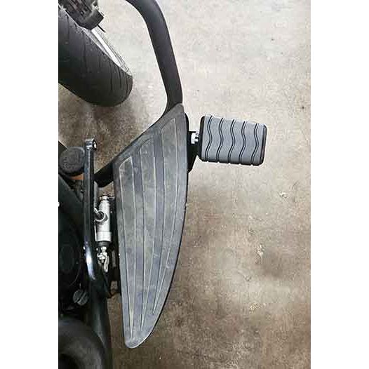 A close-up of the Adjustable Foot Rest Kit Anti Vibration Style by Witchdoctors on a concrete floor, highlighting its textured surfaces and part of a motorcycles rear tire.