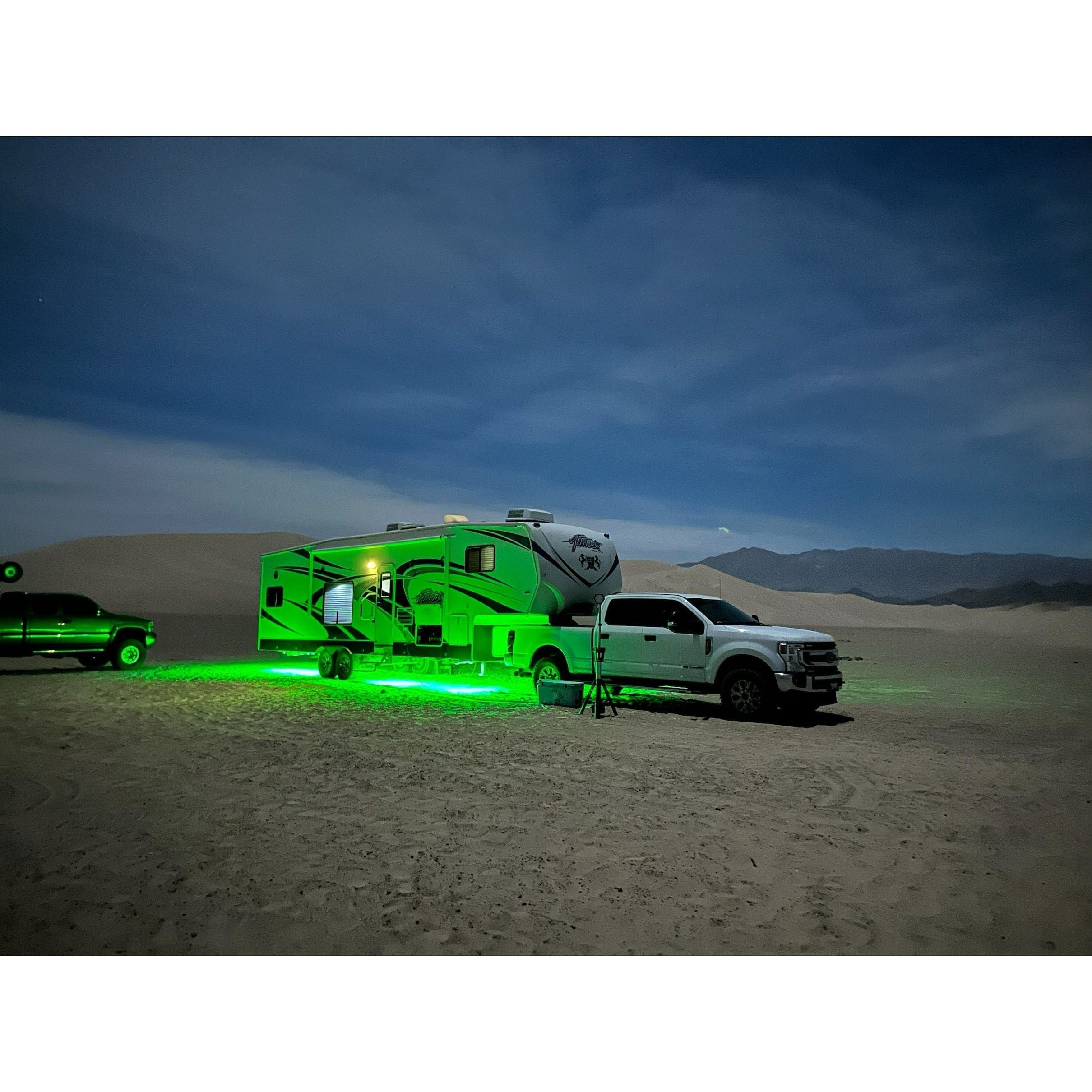 A white pickup and a green-lit RV featuring 5150 Whips RV Underglow LED Lights are parked on sand beneath a twilight sky, with dunes and distant mountains in the background. A dark vehicle is partially visible on the left.