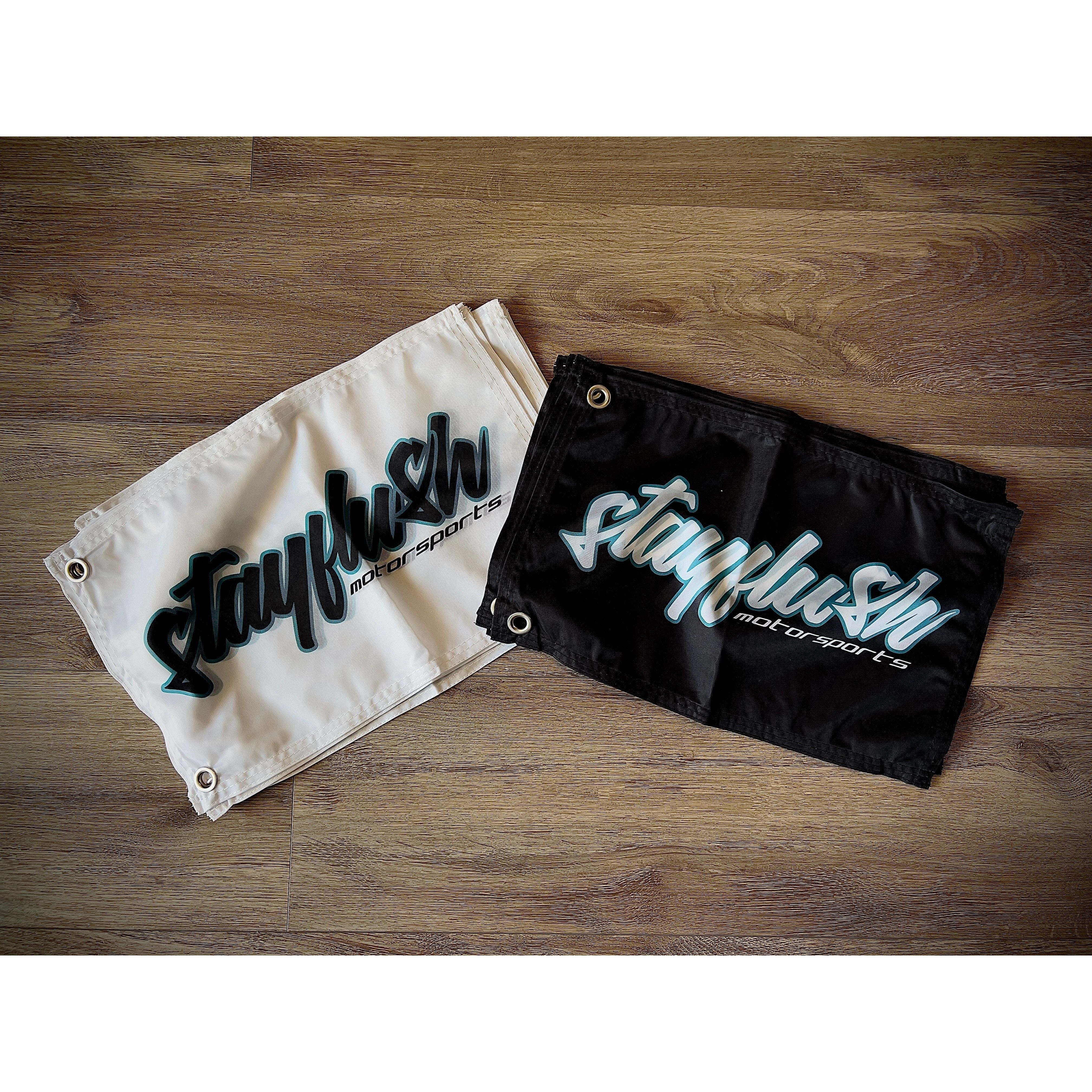 Two rectangular StayFlush 12x18 Whip Flags by StayFlush—one white, one black—lie on a wooden floor. Both feature bold “stayflush motorsports” lettering and have metal grommets in the corners for easy hanging. Single sided design.
