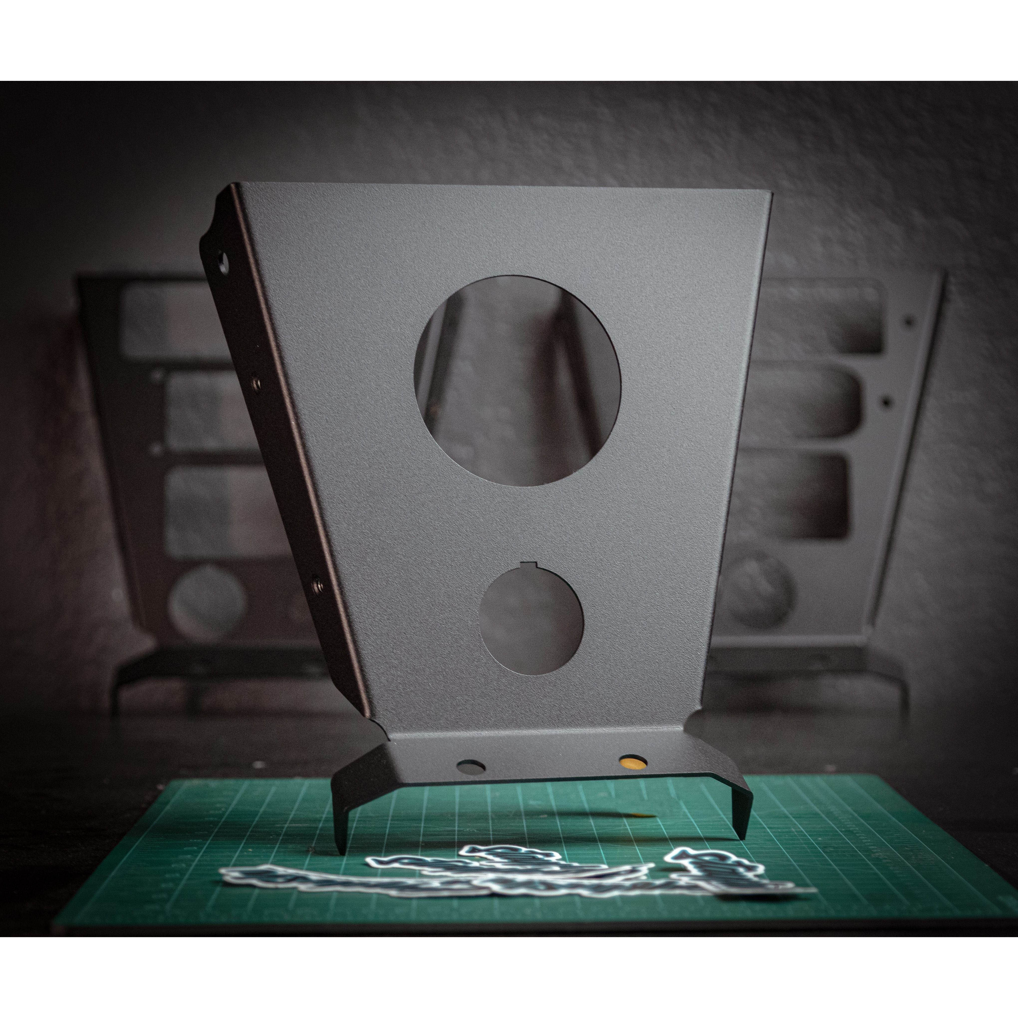 A StayFlush Cyclops Console matte gray metal bracket with circular cutouts, ideal for Canam X3 console or head unit installation, stands upright on a green cutting mat, with similar brackets blurred in the background.