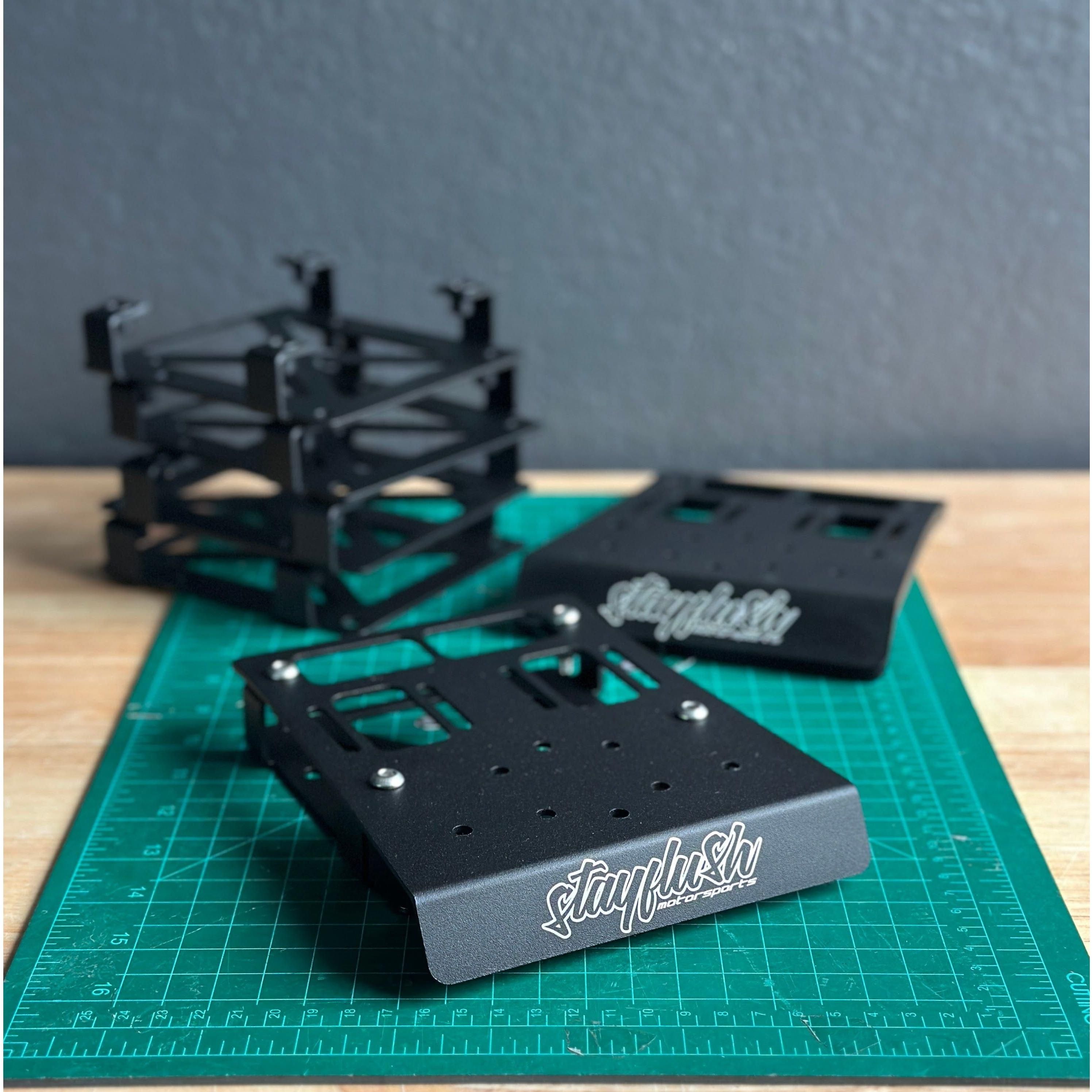 Two StayFlush Can-Am X3 Dash Device Mounts with logos, designed for AMPS pattern devices, rest on a green cutting mat. Black assembled mount components are stacked in the background against a gray wall.