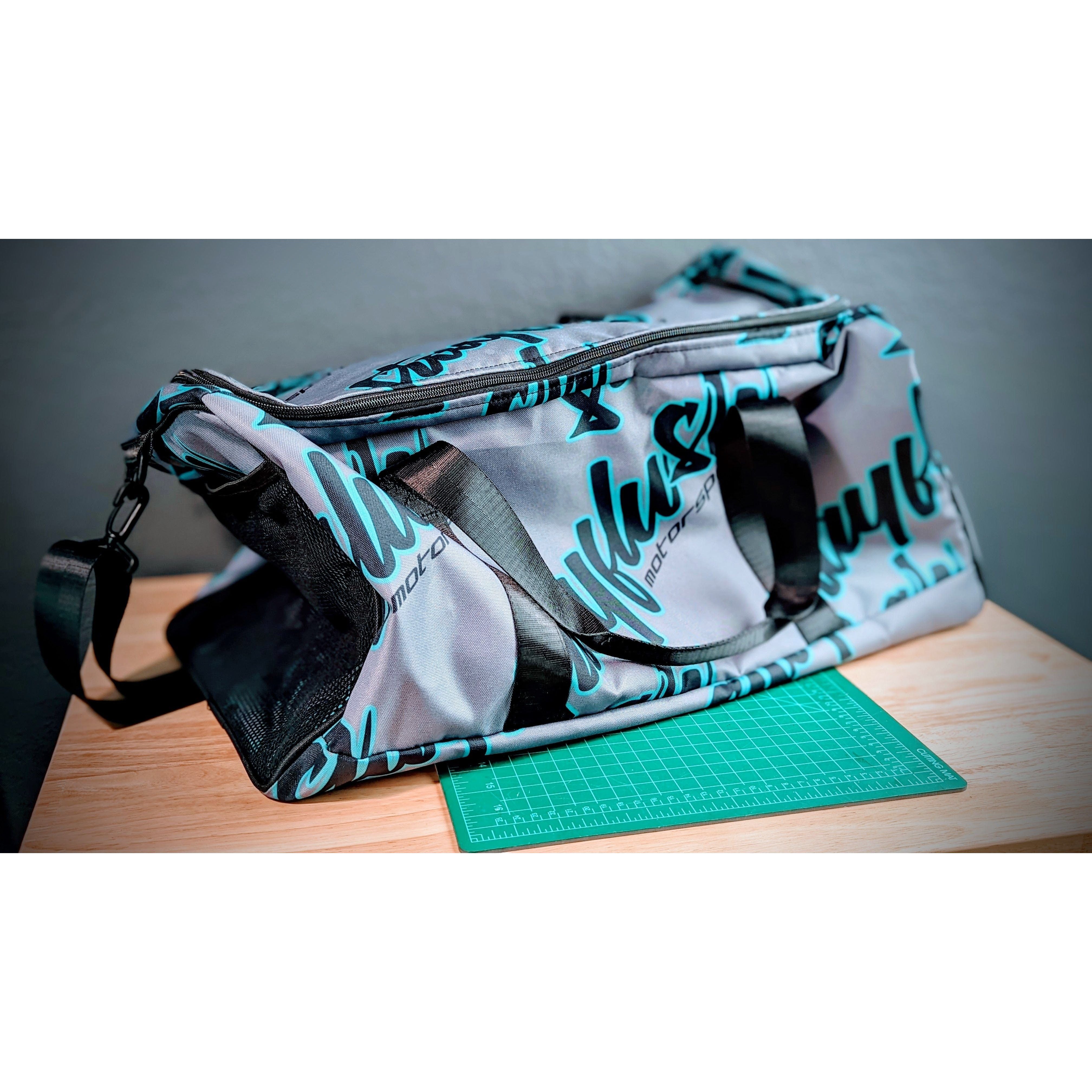A gray and teal StayFlush Duffle bag by StayFlush with black handles sits on a wooden table over a green cutting mat. Featuring bold branding and an adjustable shoulder strap, it’s ideal for carrying riding gear. The background is a plain gray wall.