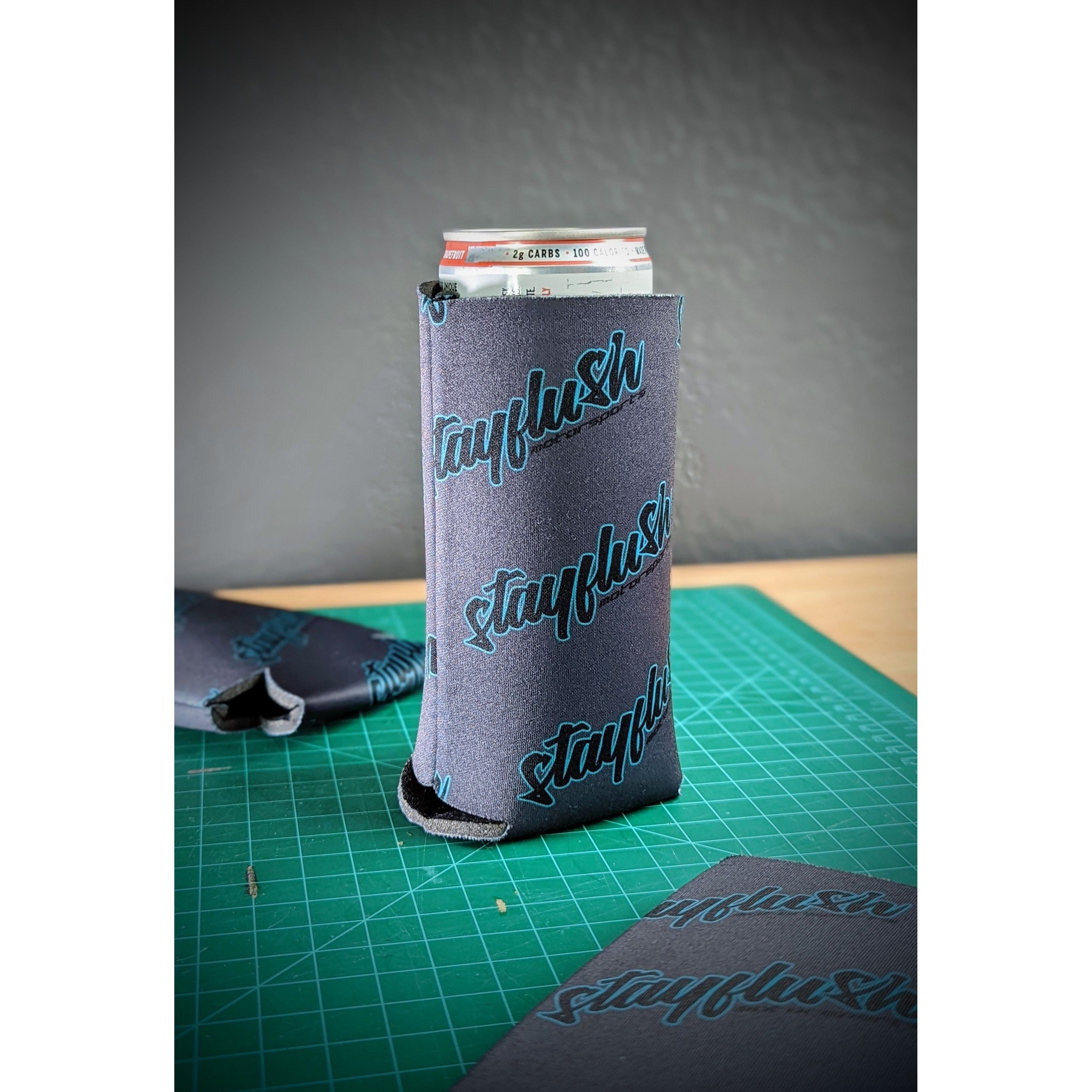 The StayFlush Coolie by StayFlush, a gray neoprene sleeve with teal StayFlush text, keeps your tall can cool in style on a green cutting mat beside another sleeve and pen, set on a wooden surface against a gray wall.