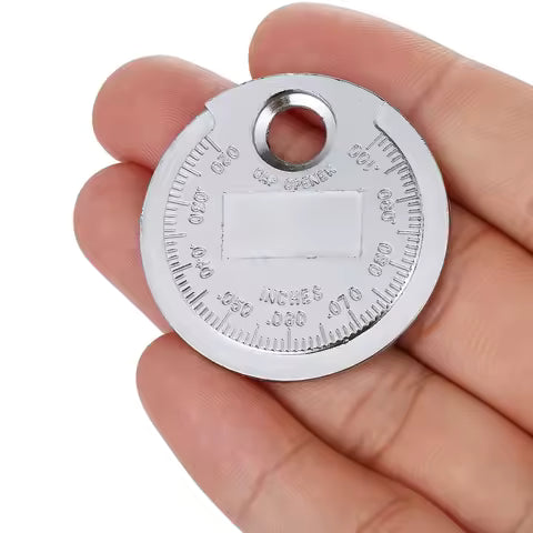 A hand holds the Spark Plug Gap Tool by Witchdoctors, a round metal tool with engraved inch and millimeter markings along the edges and a large hole near the top.