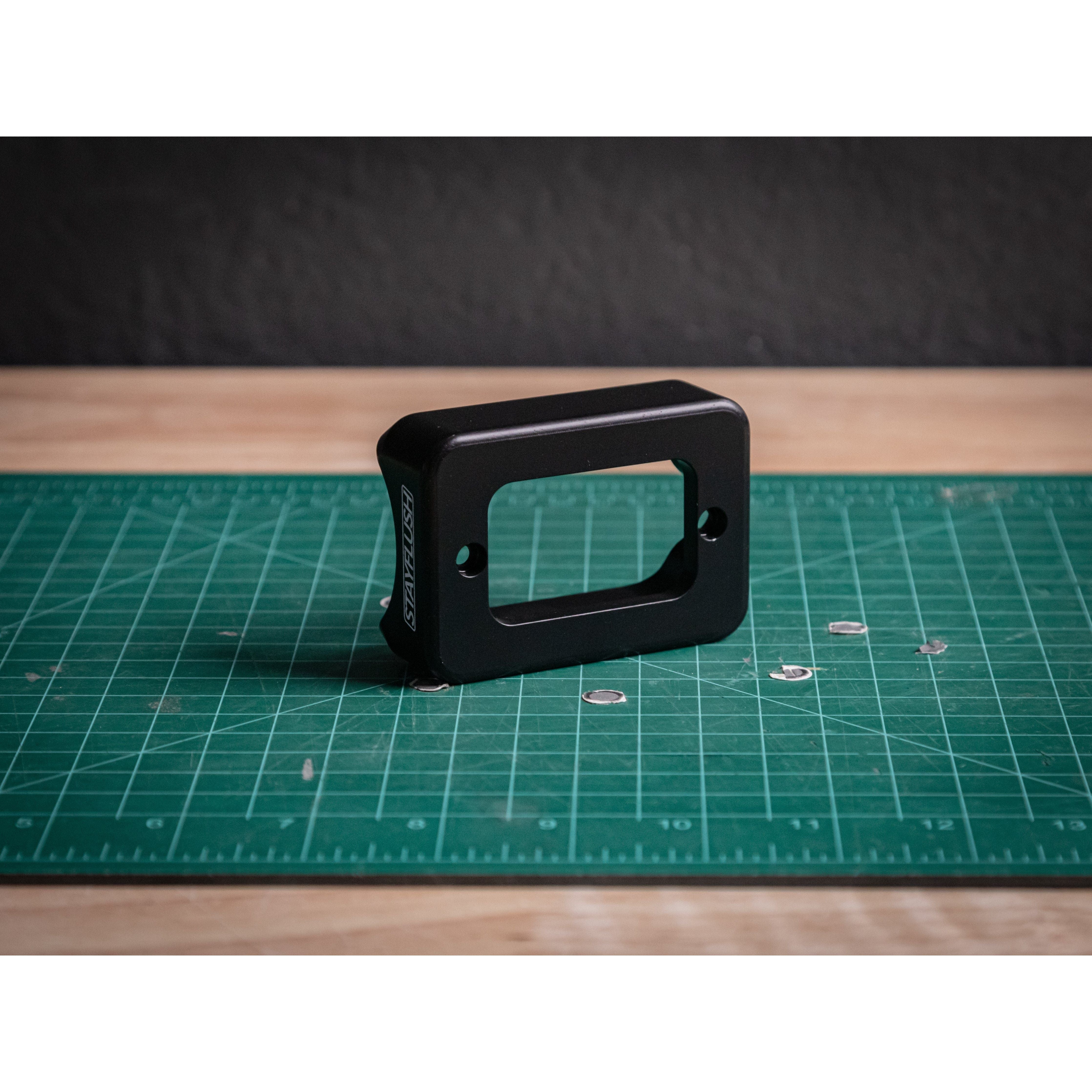 The BD Dome/Rock Light Mount by StayFlush is a black rectangular plastic bracket with two screw holes, shown upright on a green grid cutting mat—perfect for Baja Designs or off-road lighting setups.