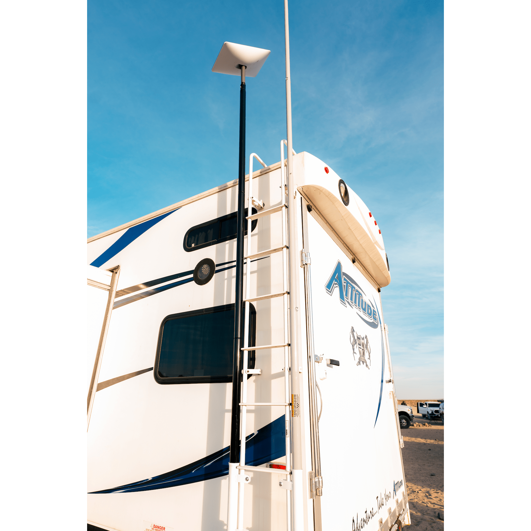 A Starlink Mount Kit by 5150 Whips is installed on a tall pole at the back of a white RV with blue accents and a ladder, set against a clear blue sky.