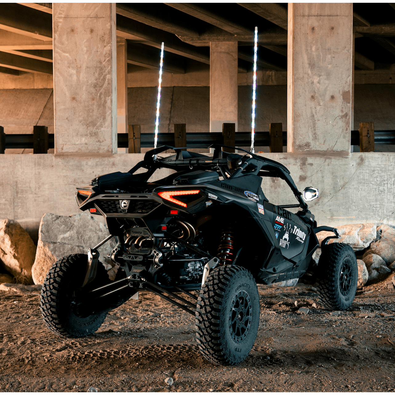 A black off-road vehicle with large rugged tires is parked under a concrete bridge on dirt and rocks, showcasing the customizable 2x 187 Chalk Line LED Whips (1 Pair) by 5150 Whips, which feature Bluetooth-enabled LED lighting.