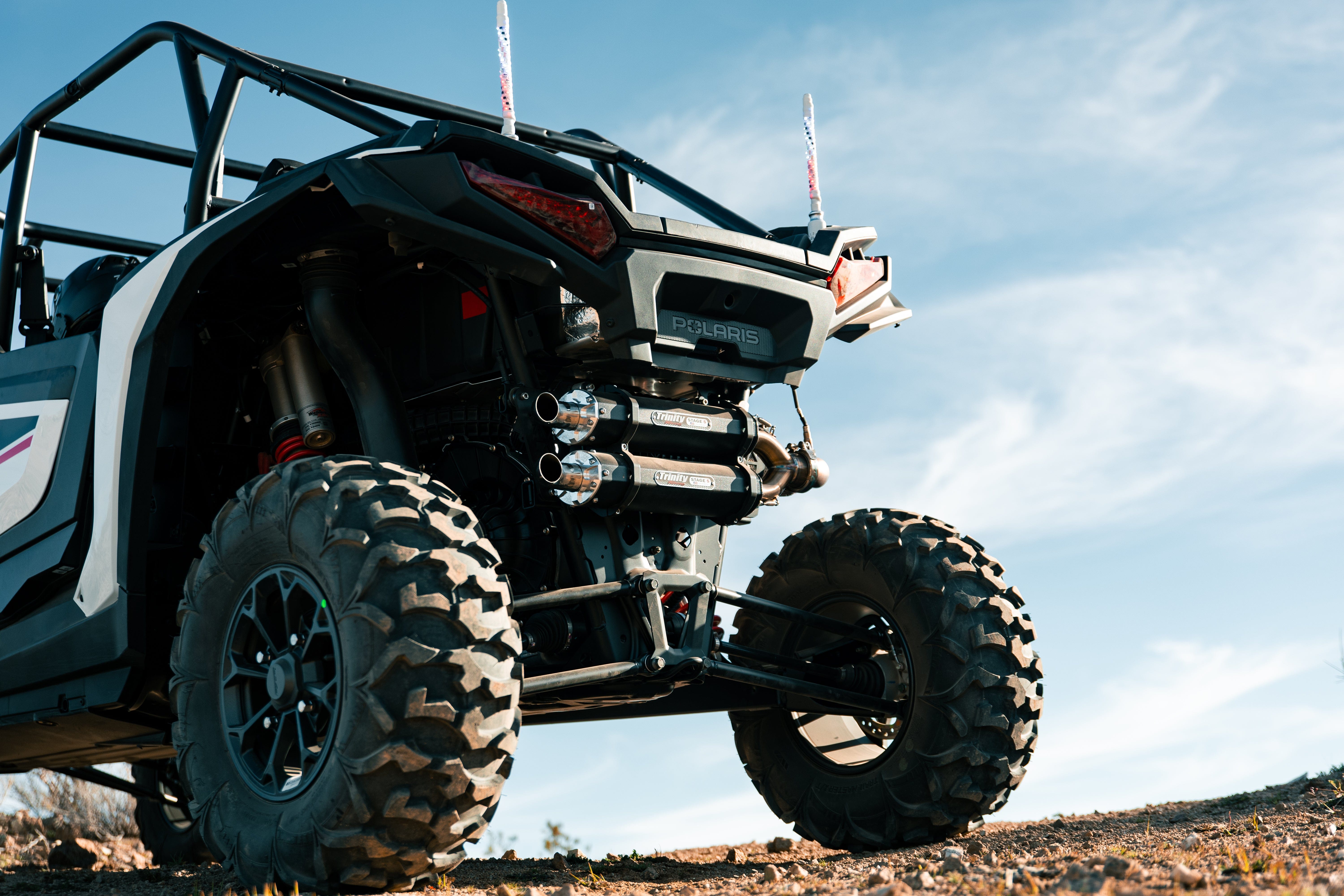A close-up, low-angle shot of a Polaris featuring the 2024+ Rzr XP 1000 Slip On by Trinity Racing, with rugged tires and stainless steel exhaust, parked on dirt under a blue sky with wispy clouds.