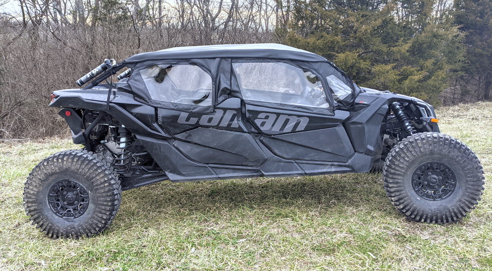 A Can-Am side-by-side with large, rugged tires and Seizmik Soft Upper Doors is parked on grass, with leafless trees in the background.
