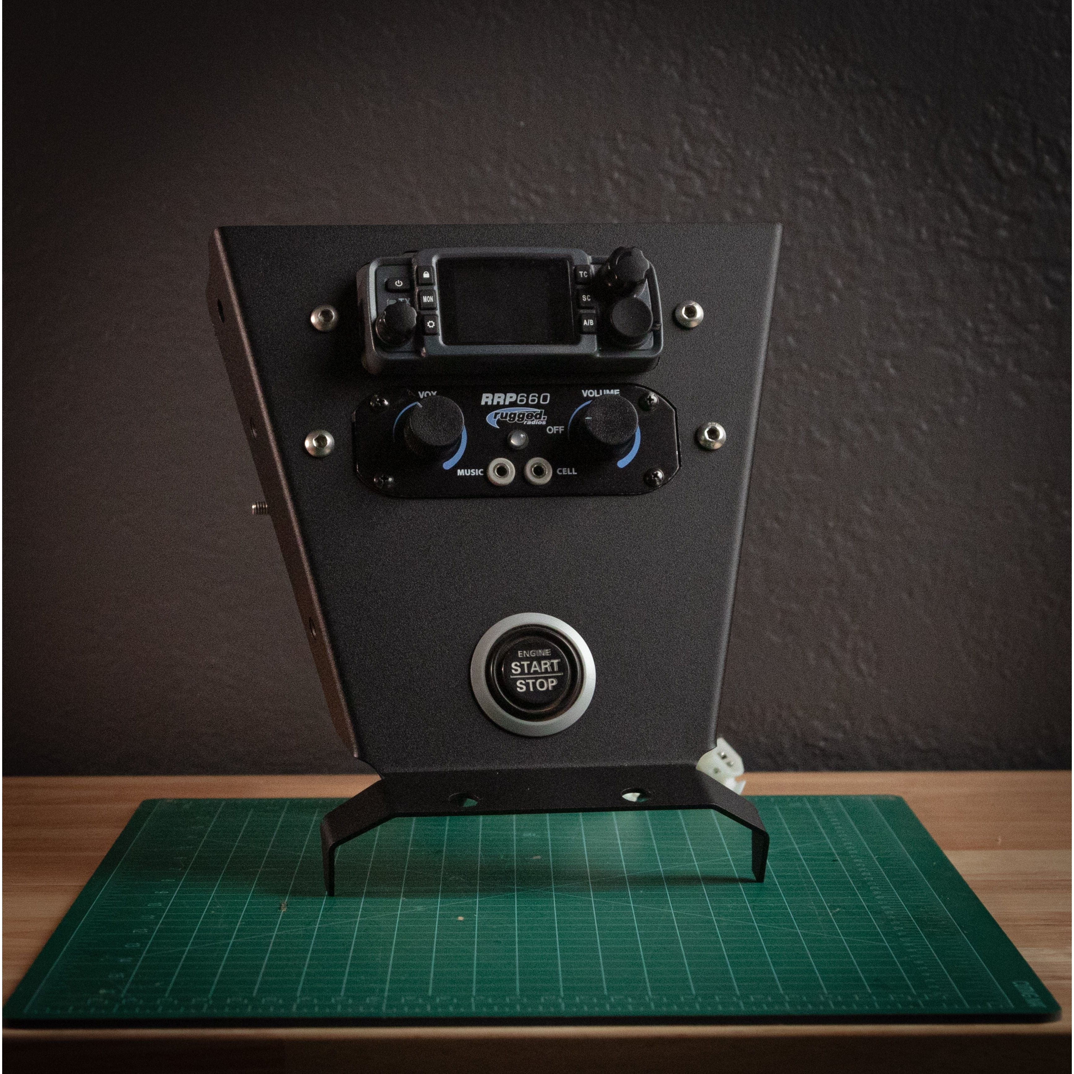 The StayFlush ABM Mini Mount (Classic) by StayFlush, a black device with knobs, digital display, and a large START/STOP button, rests on a green cutting mat beside laser-cut parts on a wood surface against a dark textured wall.