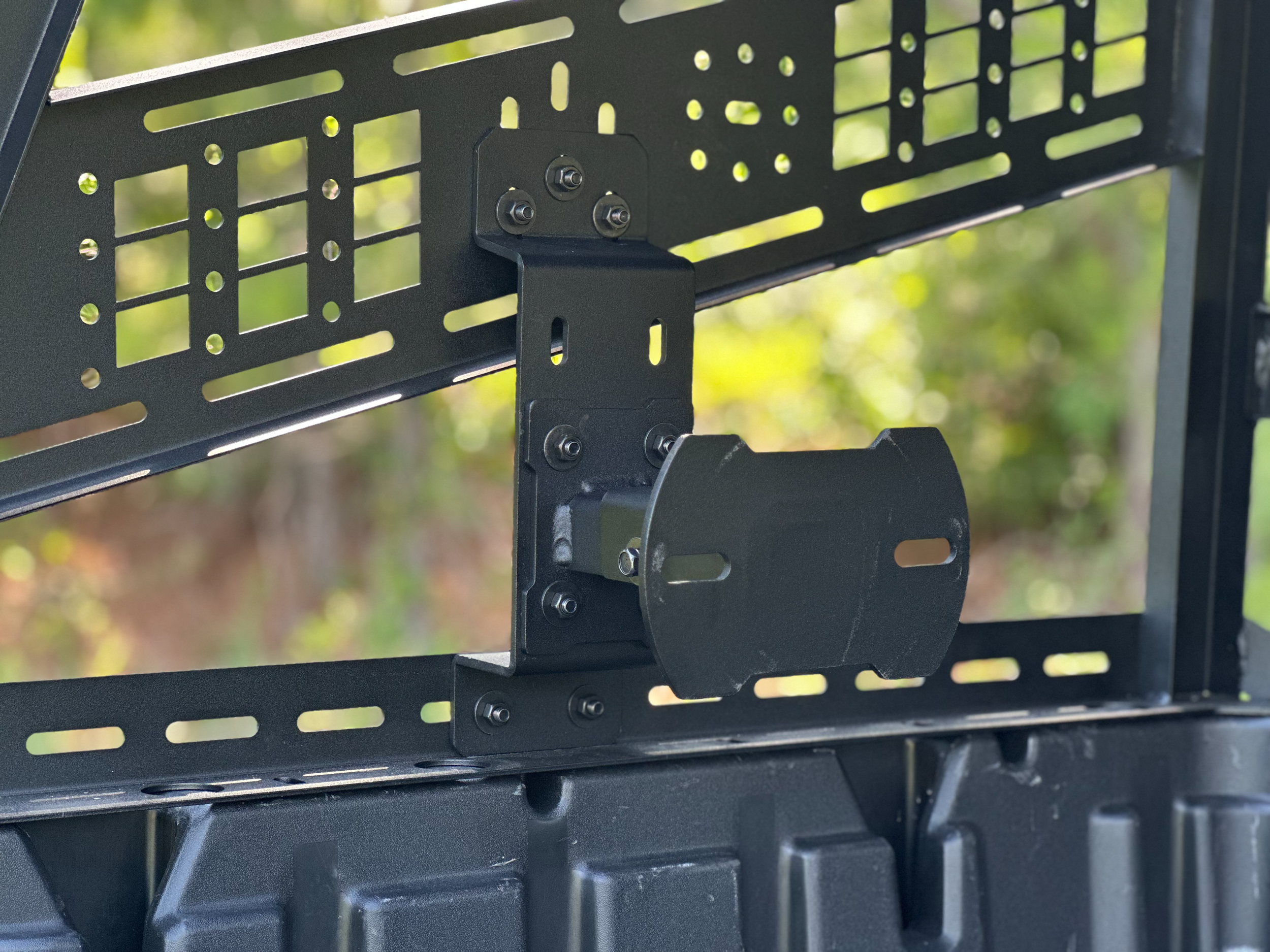 A close-up of the Kawasaki Ridge Tool-Less Versa-Vent UV Resistant Front Windshield Kit by Seizmik shows its black metal mounting bracket with multiple bolts and slots for adjustable installation, attached securely against a backdrop of blurred green foliage.