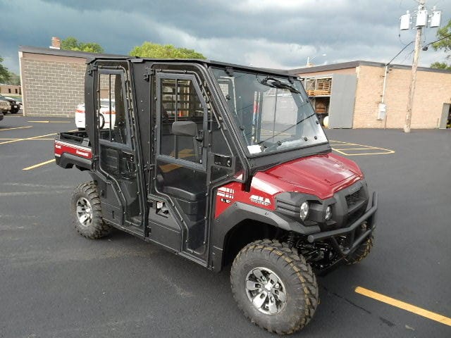 A Full UTV Cab by Open Trail, featuring red and black colors, four doors, and polycarbonate panels for visibility, is parked on a paved lot near brick buildings under a cloudy sky.