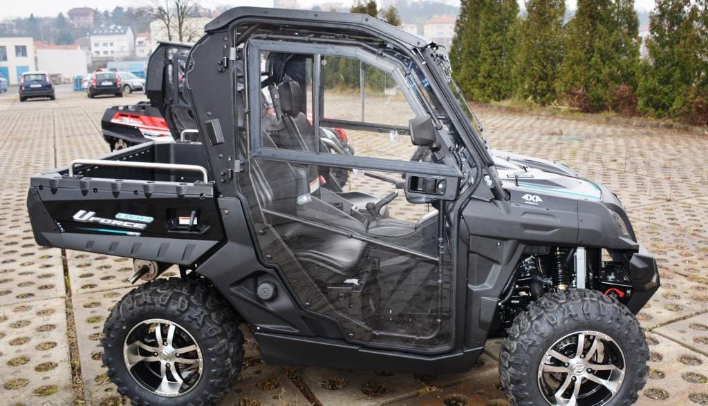 The Full UTV Cab by Open Trail, featuring large off-road tires and polycarbonate door panels, is parked outdoors on concrete with clear views of trees and buildings in the background.
