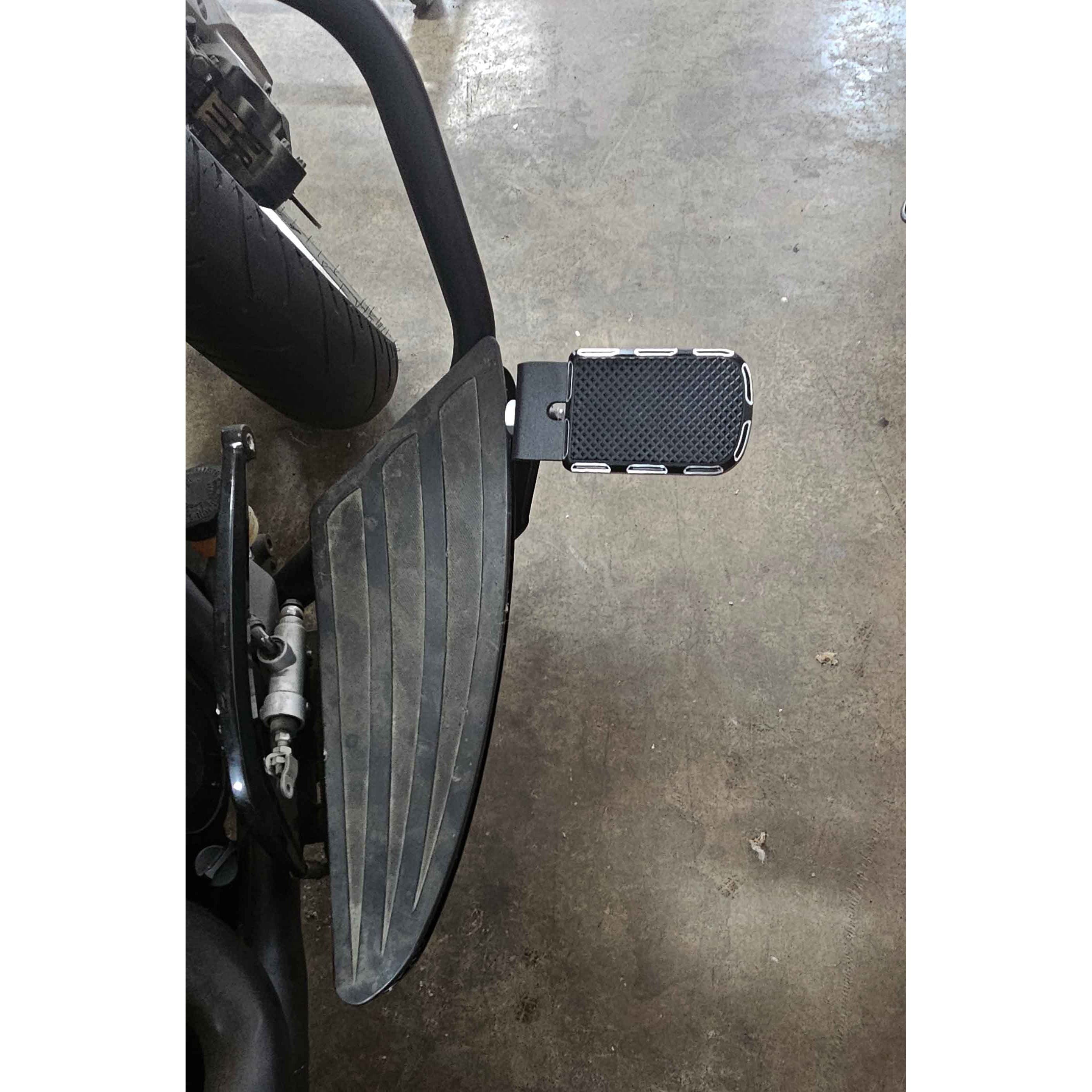 Close-up of the Adjustable Foot Rest Kit Beveled Style by Witchdoctors, featuring large grooved rubber footrests and adjustable pegs on a concrete floor. The design enhances motorcycle comfort. A rear wheel is partially visible in the background.