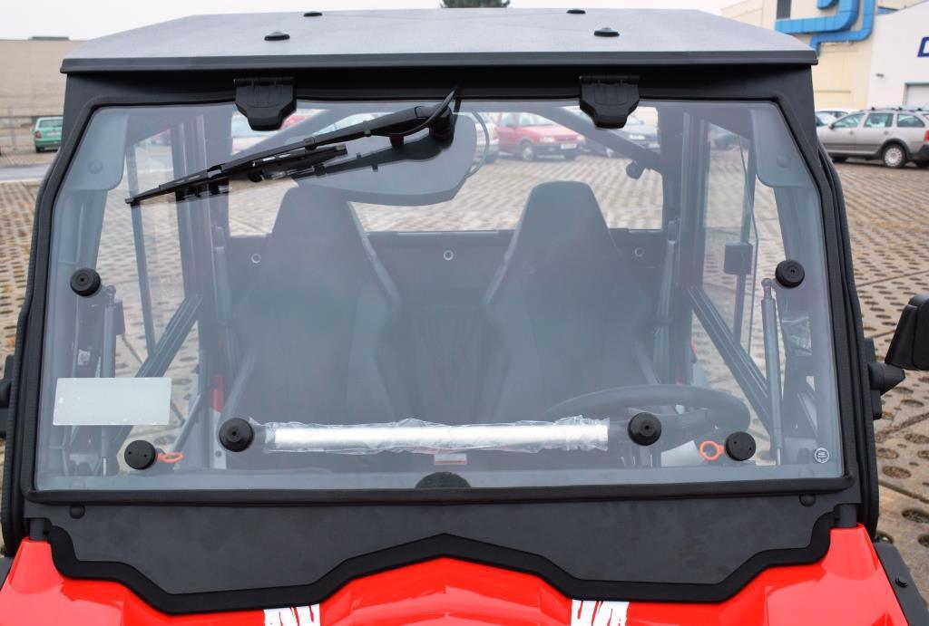 Front view of a red UTV with the Full UTV Cab by Open Trail, featuring polycarbonate door panels, clear windshield with wiper, visible seats and steering wheel, parked on pavement with buildings and cars in the background.