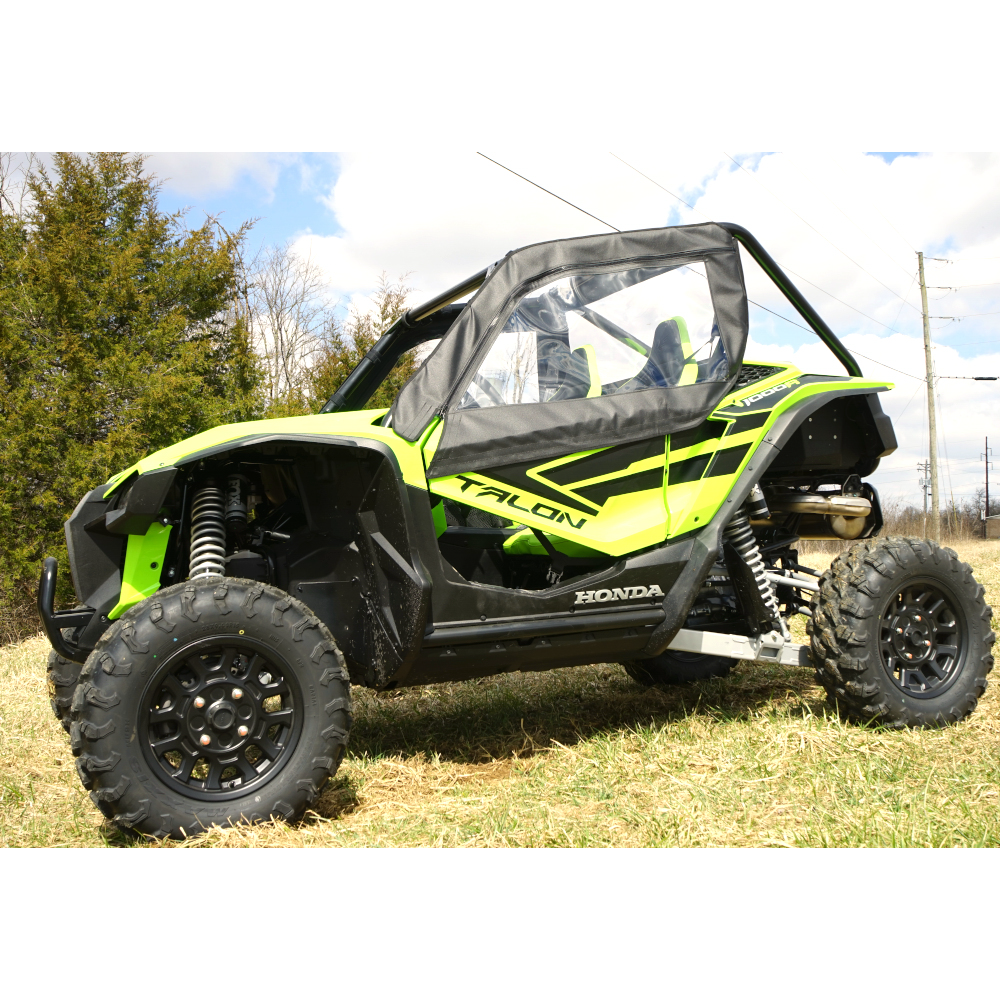 A Kawasaki Pro Fx with a Seizmik Soft Door Kit is parked on grass. The off-road vehicle features rugged tires, a roll cage, and a partially enclosed cabin. Trees and a partly cloudy sky are in the background.