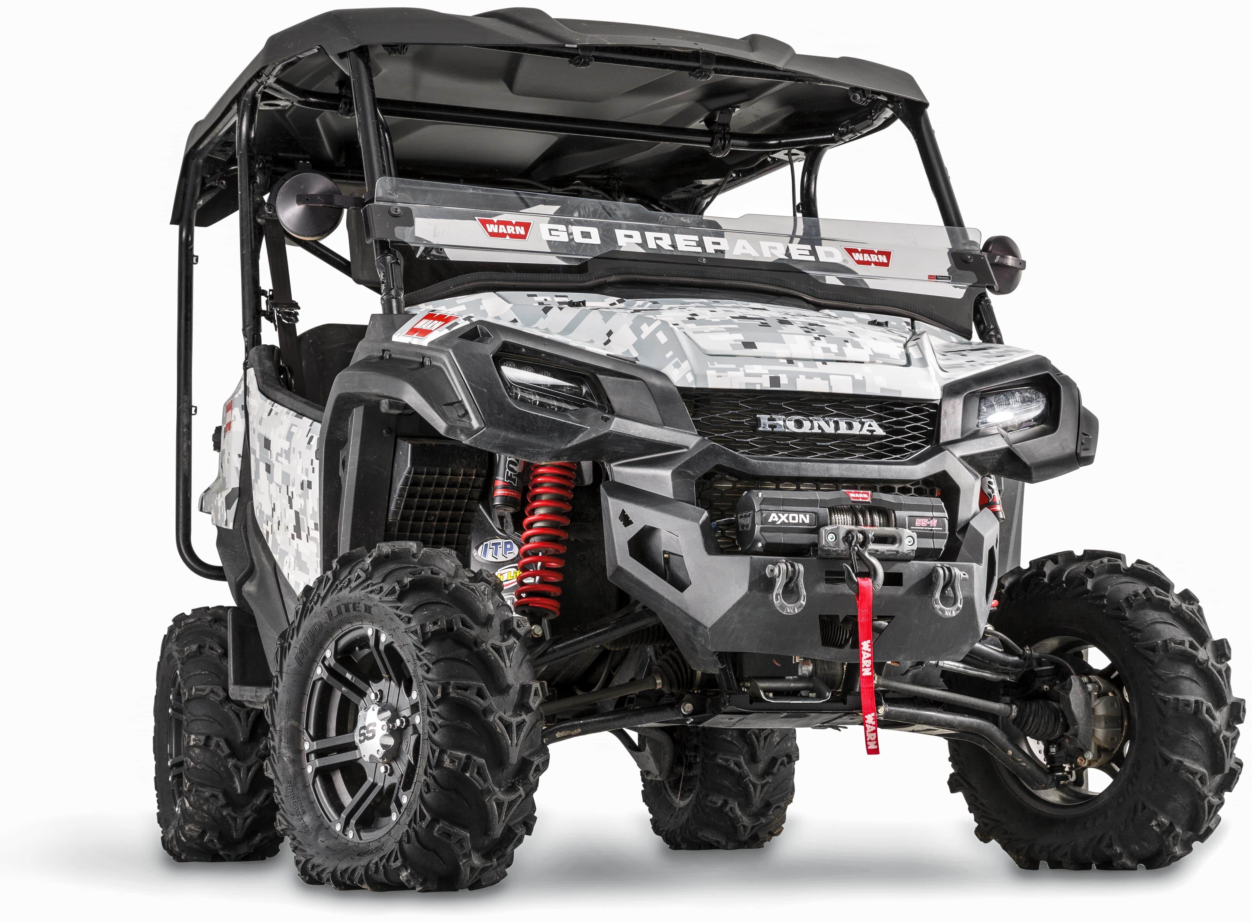 A durable steel Honda Pioneer 1000 (2016-2022) front bumper winch mounting kit by Warn, designed for rugged off-road utility vehicles with large all-terrain tires and a camo exterior, shown from a low front angle.