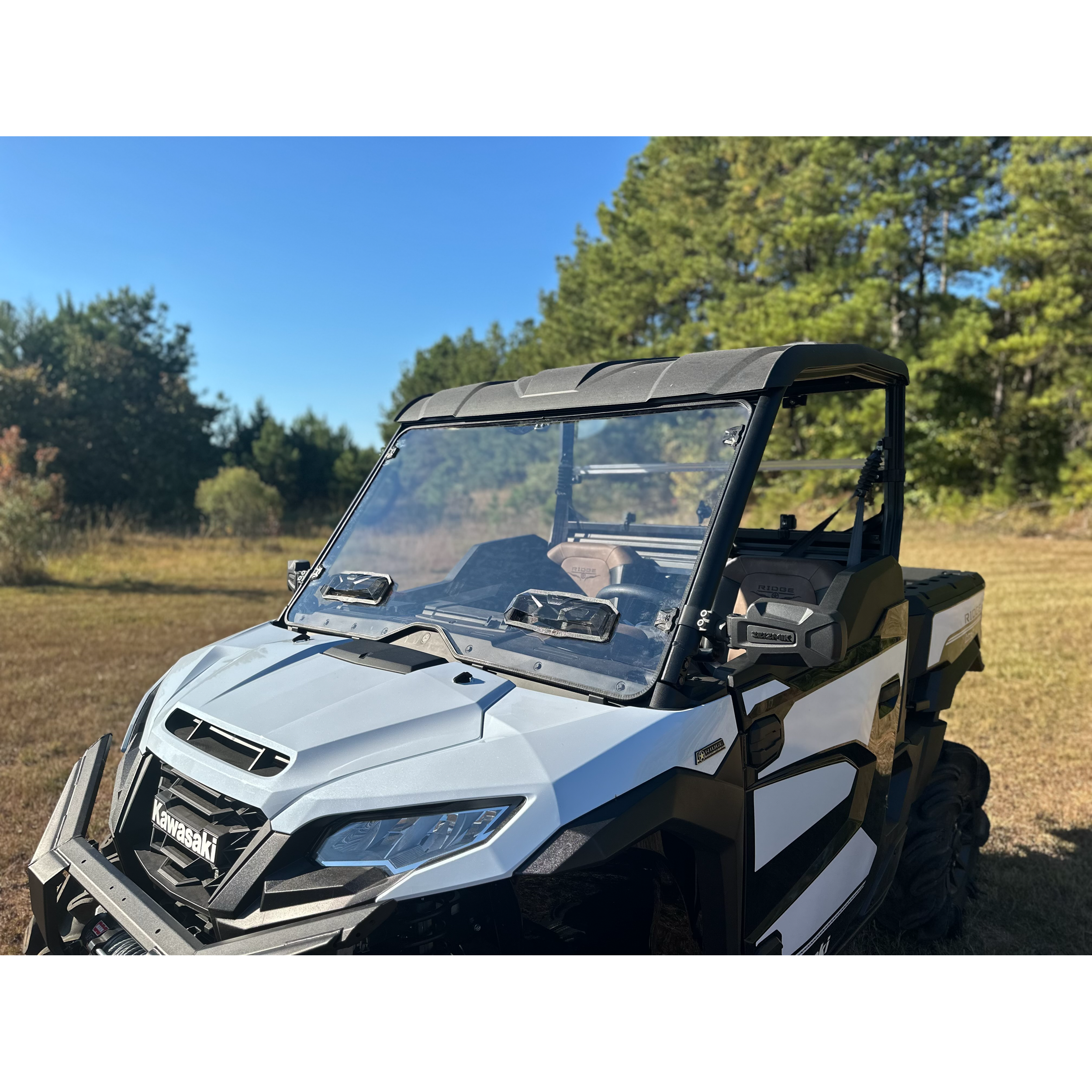A Polaris Composit Roof by Seizmik, made from heavy-duty high-density plastic, tops a white and black utility vehicle parked on dry grass near a wooded area under clear blue sky and green pine trees.