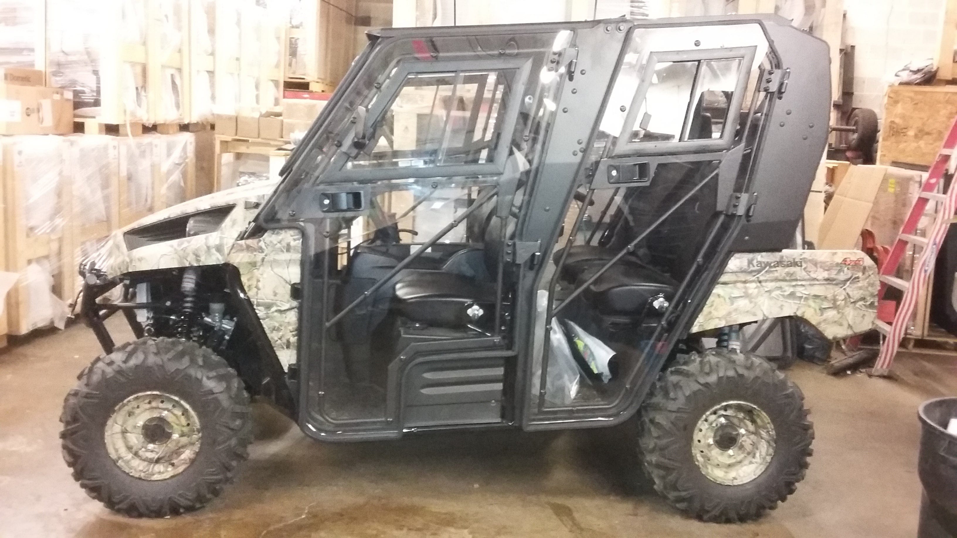 A Full UTV Cab by Open Trail, featuring a camouflage-pattern off-road vehicle with large rugged tires and transparent polycarbonate door panels, is parked indoors on a concrete floor surrounded by boxes and equipment.