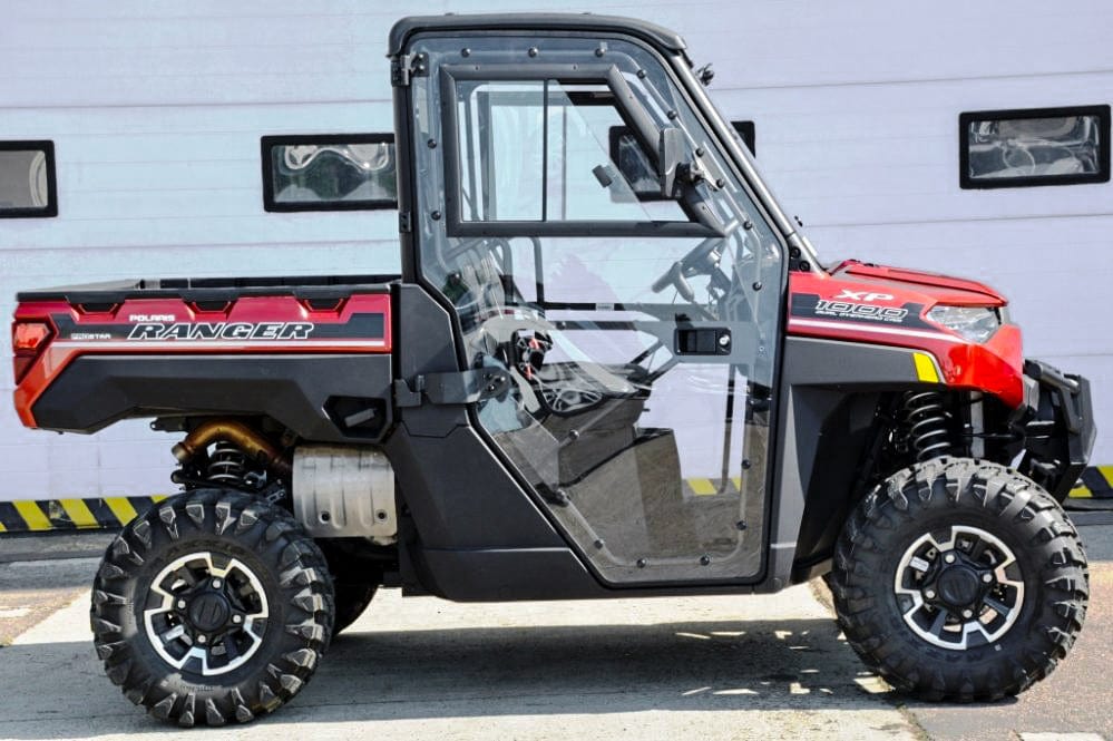 A red and black Polaris Ranger UTV with large off-road tires and a Full UTV Cab by Open Trail featuring polycarbonate door panels is parked in front of a white garage door, offering rugged construction and clear doors for versatile outdoor use.