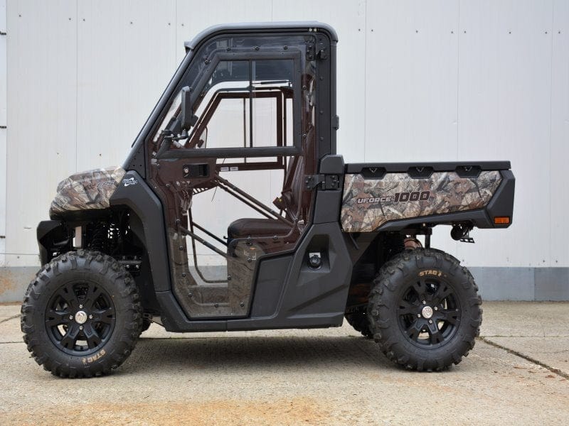 Side view of the Full UTV Cab by Open Trail, featuring large off-road tires, clear polycarbonate door panels, and a rugged design, parked on concrete before a plain white wall.
