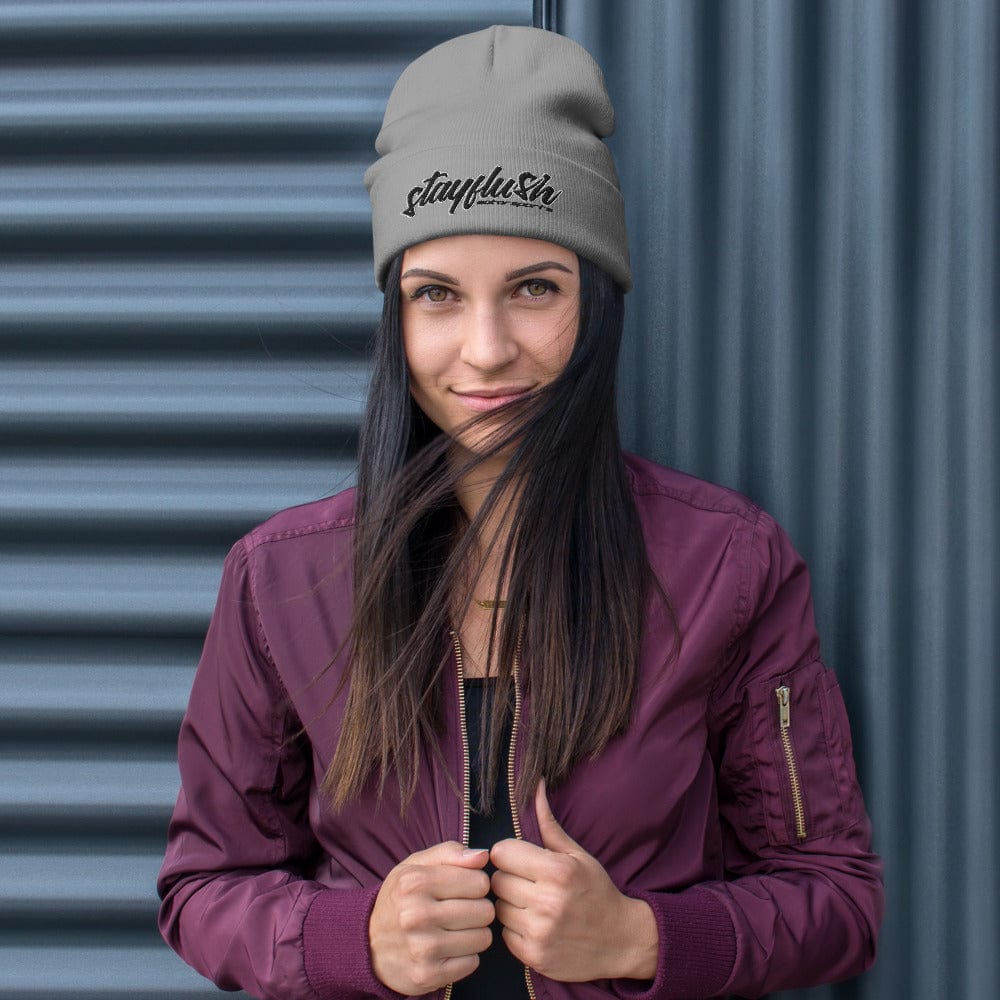 A woman in a maroon jacket and gray StayFlush Beanie by StayFlush, featuring “stayplusln” text, stands before a corrugated metal wall with a slight smile as her long hair blows across her face. The breathable cotton blend offers comfort and fits most.