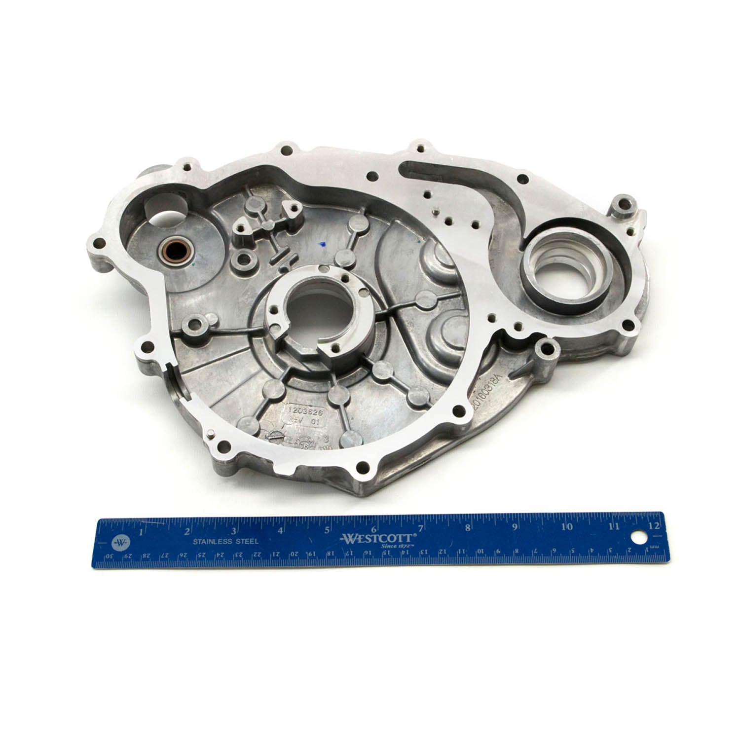 Silver metal stator cover with multiple mounting holes and circular recesses, placed above a blue ruler for scale.
