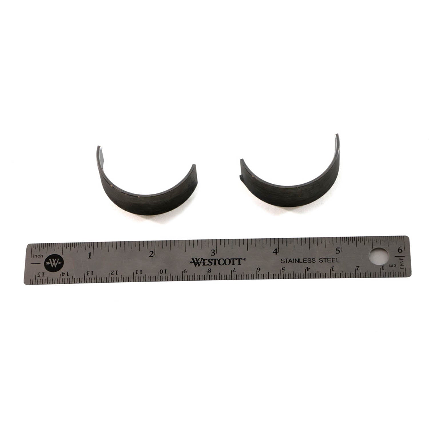 Two curved metal crankpin components with a gray finish positioned above a metal ruler showing inches.