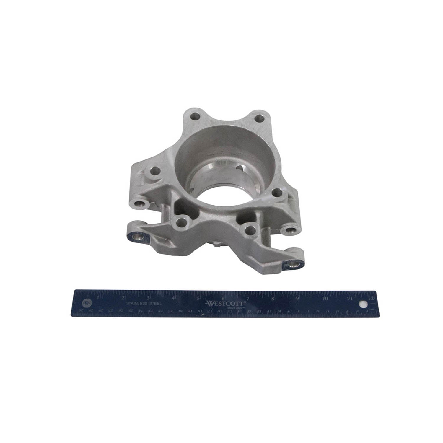 Silver metal steering knuckle with mounting holes and circular hub, placed above a blue ruler for scale.